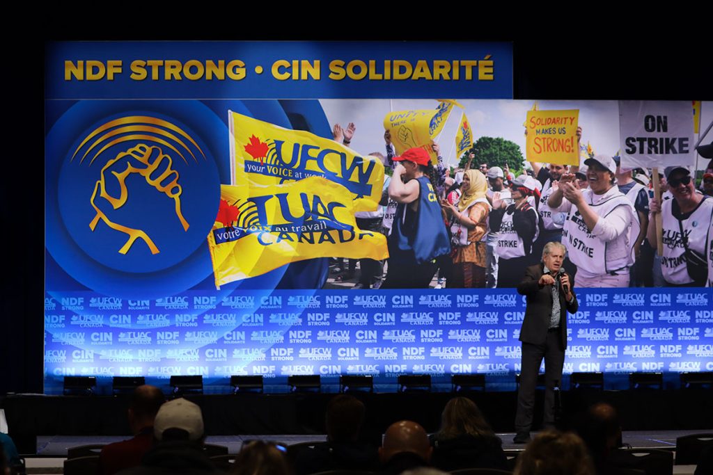 Charlie Angus speaks into a handheld mic standing on stage.