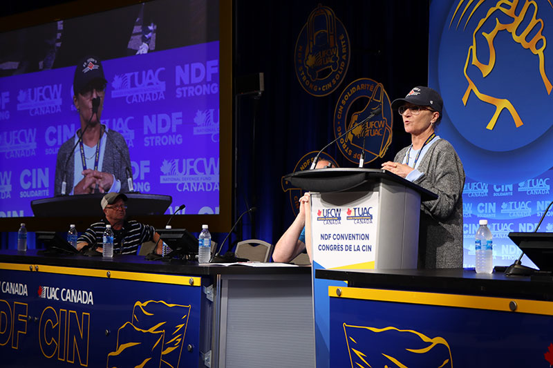 A UFCW member speaks at the podium on stage and can be seen on a large display screen in the background. Another person is seated on stage at a table.