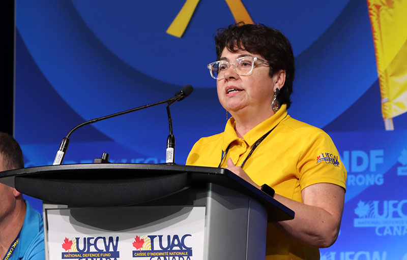 A person in a gold UFCW shirt stands at a podium speaking into a microphone at the 2025 NDF Convention.