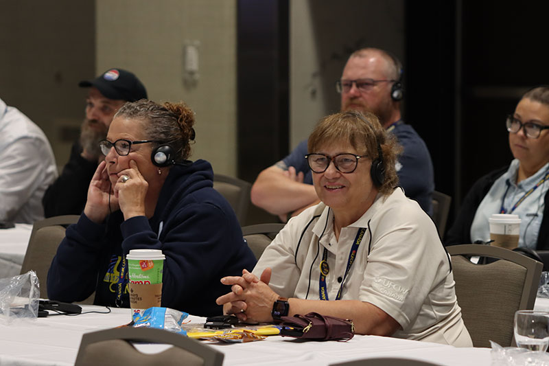 Two people seated in the convention audience who are both wearing translation devices. One is smiling. Others are seen seated in the background.