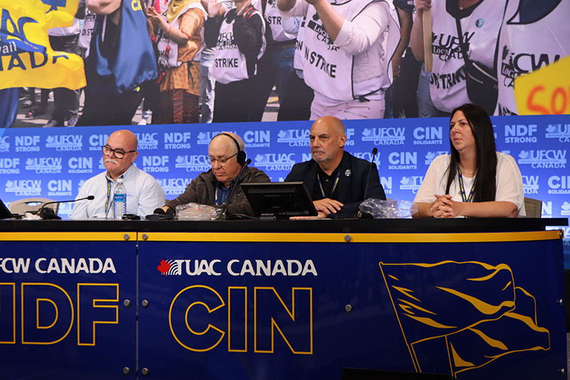 Four members of the NDF Committee sit on stage at a table with UFCW Canada NDF logos.