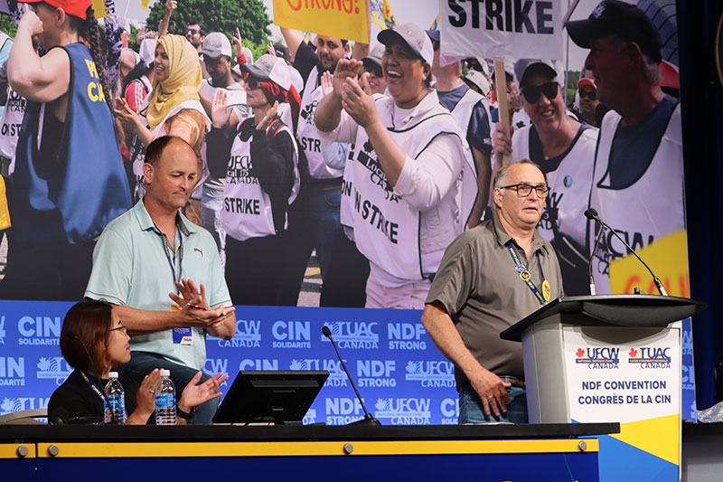 Two people on stage applaud a third person who is speaking at the microphone. A large banner with images of workers on strike is in the background.