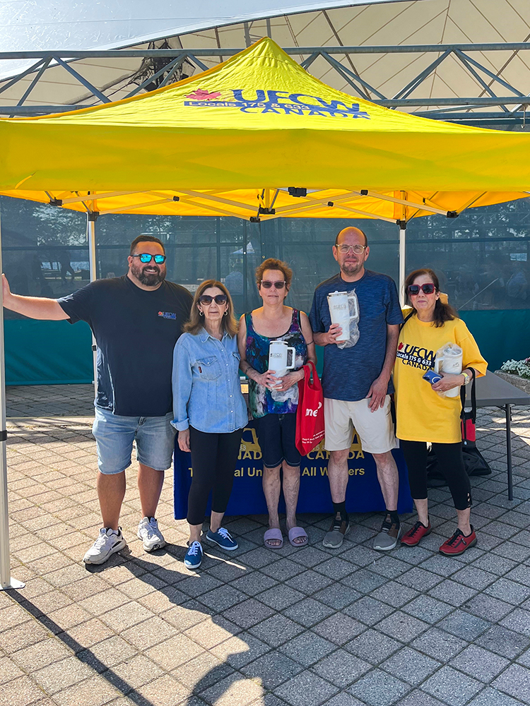 Five people stand under a yellow UFCW tent. Three people are holding large insulated tumbler mugs at the Sault Ste. Marie Labour Day 2025 event.