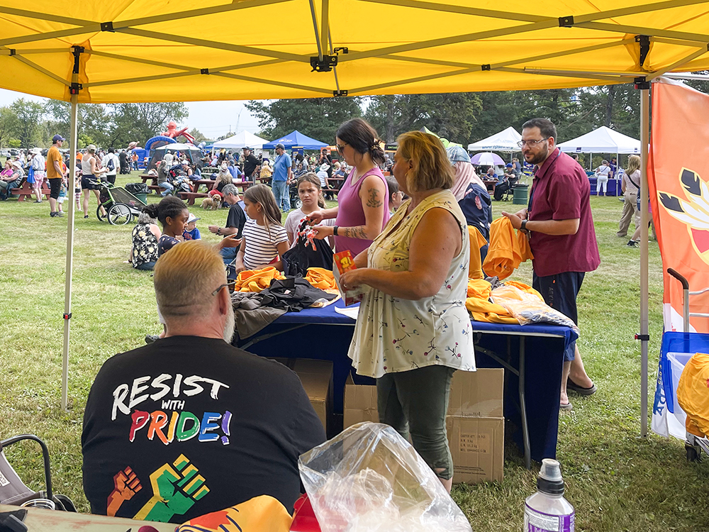 UFCW staff greet members and guests at a yellow UFCW tent at the Thunder Bay picnic. Many more people can be seen walking behind among other tents.
