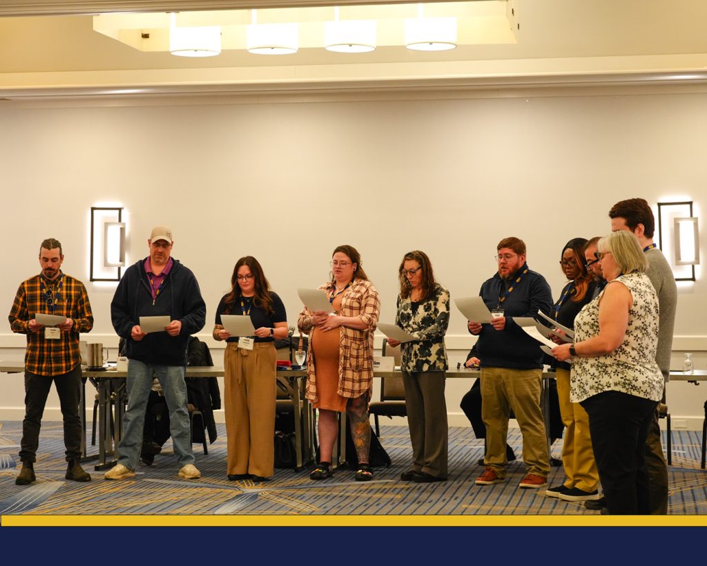 10 people stand, holding pieces of paper, reading