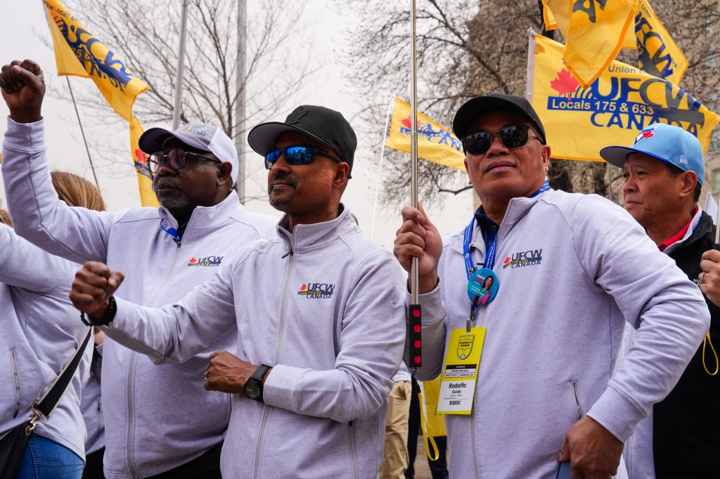 3 UFCW delegates wearing UFCW jackets and holding flags at a rally