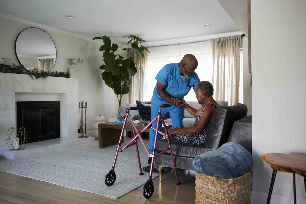 A healthcare worker helps a senior lady get up from her couch to get to her walker.