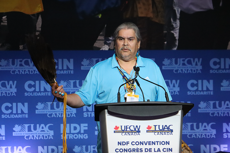 UFCW Canada Resident Elder Eric Flett holds a large black feather and speaks from the podium as he opens the 2026 NDF Convention