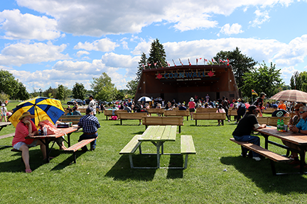 a person sits at a picnic table under a UFCW umbrella with the Cornwall bandstand and many people in the background