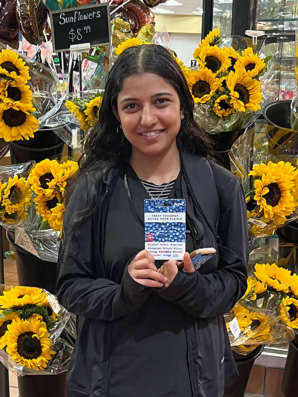 Rupinder Kaur holds a gift card while standing in front of sunflower display in a grocery store